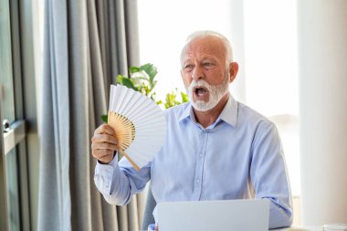 İyi olmayan kıdemli bir iş adamı dizüstü bilgisayarda el pervanesiyle oturuyor. Aşırı ısınmış erkek işçi, ofiste sıcak çarpmasından ve iş yerinde klimasız kalmaktan dolayı bocaladı..