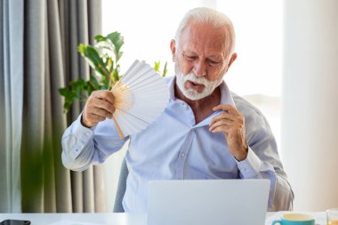 İyi olmayan kıdemli bir iş adamı dizüstü bilgisayarda el pervanesiyle oturuyor. Aşırı ısınmış erkek işçi, ofiste sıcak çarpmasından ve iş yerinde klimasız kalmaktan dolayı bocaladı..