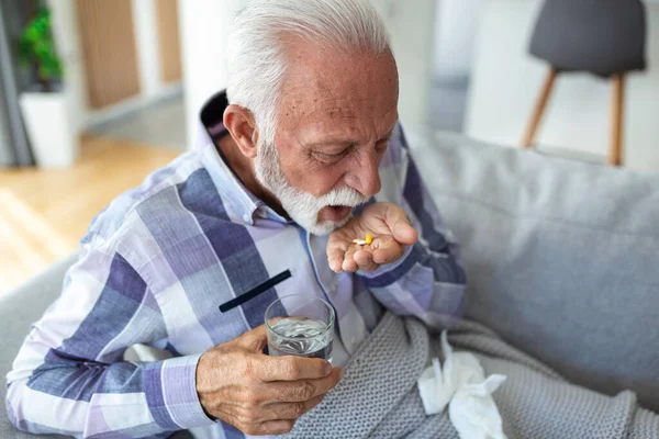 Senior man taking pills while sitting on couch at home, holding white jar with treatment, copy space. Grey-haired elderly man using supplements or vitamins. Healthy lifestyle in senior age concept