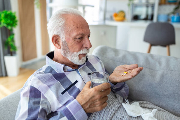 Senior man taking pills while sitting on couch at home, holding white jar with treatment, copy space. Grey-haired elderly man using supplements or vitamins. Healthy lifestyle in senior age concept