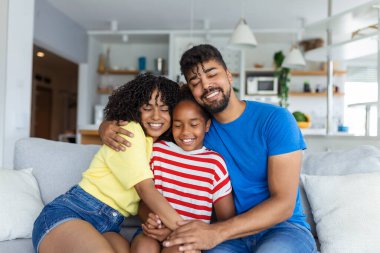 Oturma odasındaki kanepede oturan çok ırklı aile hafta sonları birbirleriyle iletişim kurarak boş vakitlerin tadını çıkarıyor. Güzel kız, anneyi kucaklıyor. Anne ve babasıyla içeride eğleniyor.