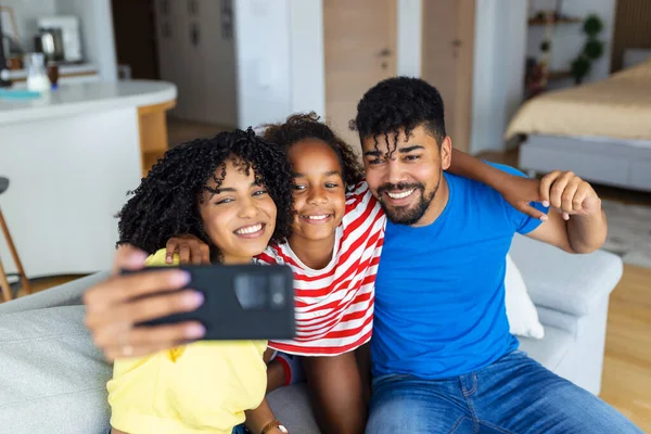 Happy African couple of parents and laughing daughter resting on couch holding smartphone, taking selfie on cellphone, making video call, looking at mobile phone screen, smiling, giggling. Family leisure