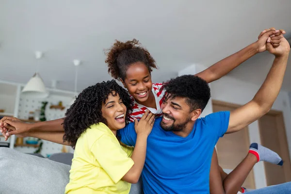Happy African American dad and mom piggybacking excited proud daughter kid, playing flying superhero, reaching arm forward. Cheerful girl riding fathers back playing active game with family at home