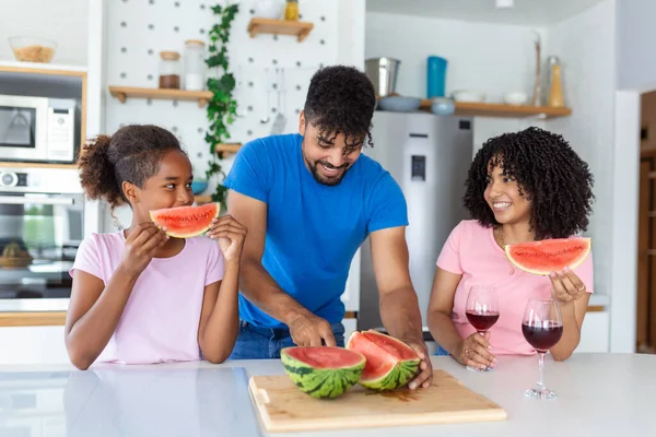 Dad is cutting watermelon while mom and daughter are watching him.