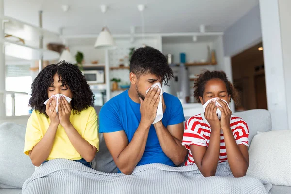 Flu And Illness Concept. Portrait of sick young African American family blowing noses with napkins together while sitting on the couch covered with blanket. Four unwell people with cold at home