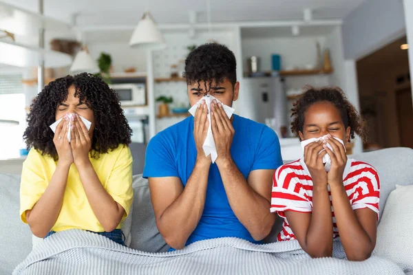 Portrait of sick young black family of three people blowing runny noses while sitting together on the sofa with napkins and covered with blanket.