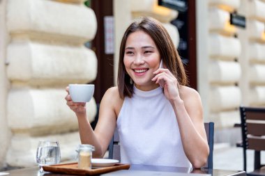 Gülümseyen Asyalı kadın kahve içiyor ve cep telefonunu kullanıyor. Tatmin olmuş bir kadın kahve içiyor. Kahve dükkanında beyaz bir fincandan kahve içen güzel bir kızın portresi.
