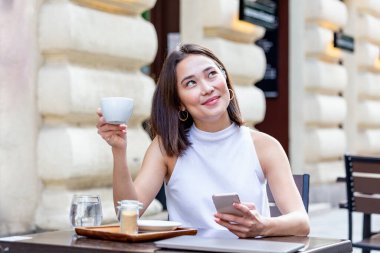 Gülümseyen Asyalı kadın kahve içiyor ve cep telefonunu kullanıyor. Tatmin olmuş bir kadın kahve içiyor. Kahve dükkanında beyaz bir fincandan kahve içen güzel bir kızın portresi.