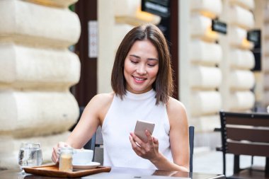 Gülümseyen Asyalı kadın kahve içiyor ve cep telefonunu kullanıyor. Tatmin olmuş bir kadın kahve içiyor. Kahve dükkanında beyaz bir fincandan kahve içen güzel bir kızın portresi.