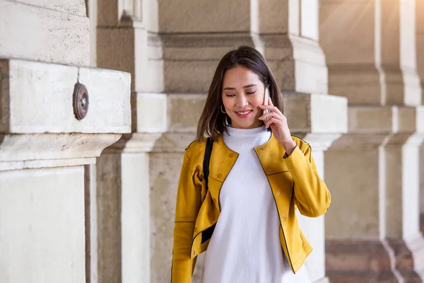Portrait of a young, attractive and beautiful Asian woman smiling as she talks on her smartphone int he city. She is looking away as she talks animatedly and happily on her mobile device.