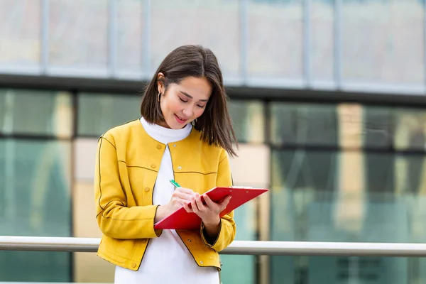 Asian Businesswoman in casual wear writing on clipboard. business woman holding clipboard. Modern, hardworking woman holding clip board in hands, writing on documents.