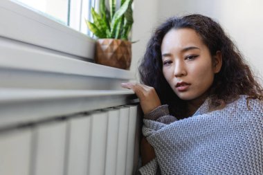 Unwell Asian woman renter in blanket sit in cold living room hand on old radiator.suffer from lack of heat . Unhealthy young woman struggle from chill freeze at home. No heating concept.