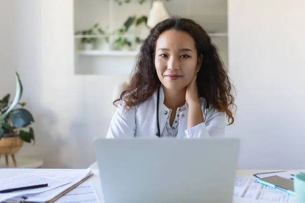Youn Asian doctor using laptop and sitting at desk. Woman professional medic physician wearing white coat and stethoscope working on computer at workplace.