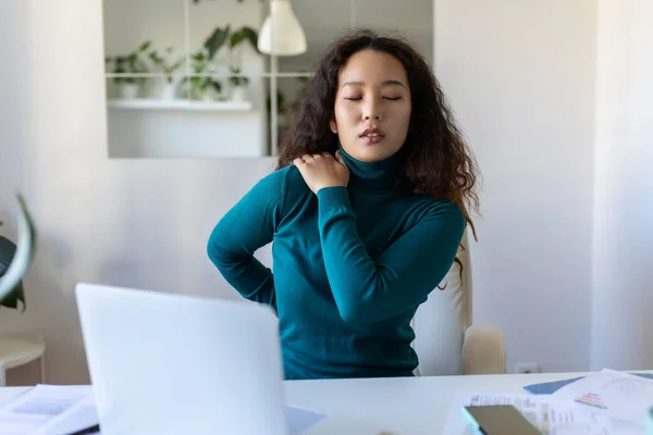 Portrait of young stressed Asian woman sitting at home office desk in front of laptop, touching aching back with pained expression, suffering from backache after working on laptop