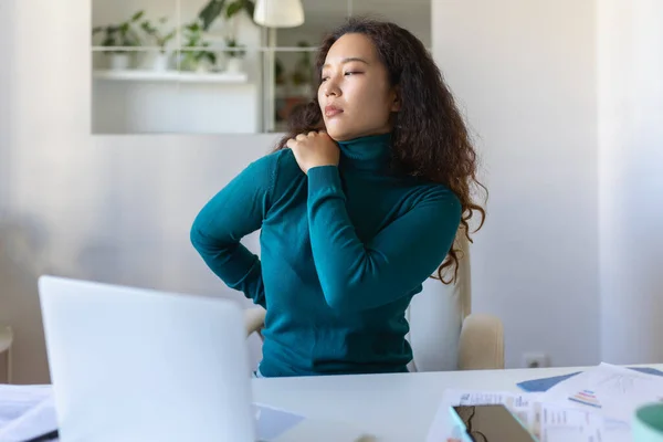 Portrait of young stressed Asian woman sitting at home office desk in front of laptop, touching aching shoulder with pained expression, suffering from shoulder ache after working on laptop