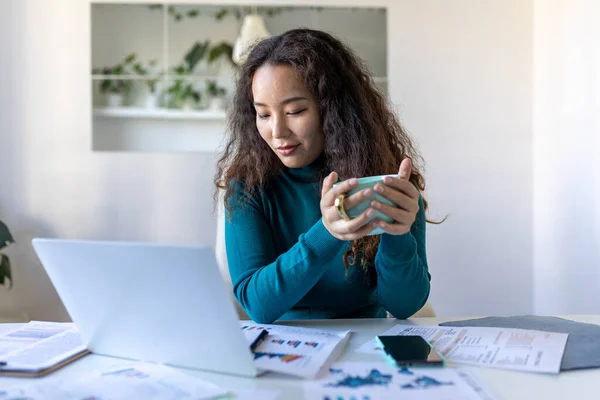 Asian woman working on laptop computer, smiling. Woman Working From Home On Laptop In Modern Apartment. Trendy woman working on laptop and drinking coffee