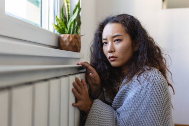 Unwell Asian woman renter in blanket sit in cold living room hand on old radiator.suffer from lack of heat . Unhealthy young woman struggle from chill freeze at home. No heating concept.