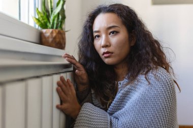 Unwell Asian woman renter in blanket sit in cold living room hand on old radiator.suffer from lack of heat . Unhealthy young woman struggle from chill freeze at home. No heating concept.