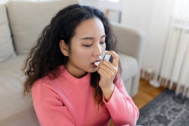 Asian woman using inhaler while suffering from asthma at home. Young woman using asthma inhaler. Close-up of a young Asian woman using asthma inhaler at home.