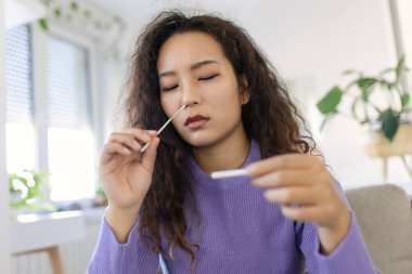 Asian woman using cotton swab while doing coronavirus PCR test. Woman takes coronavirus sample from her nose at home. woman at home using a nasal swab for COVID-19.