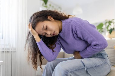 Asian woman in painful expression holding hands against belly suffering menstrual period pain, lying sad on home bed, having tummy cramp in female health concept