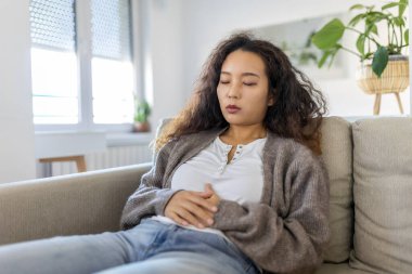 Asian woman lying on sofa looking sick in the living room. Beautiful young woman lying on bed and holding hands on her stomach. Woman having painful stomachache on bed, Menstrual period