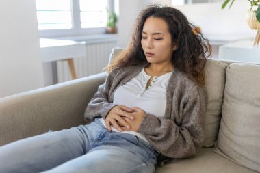 Asian woman lying on sofa looking sick in the living room. Beautiful young woman lying on bed and holding hands on her stomach. Woman having painful stomachache on bed, Menstrual period