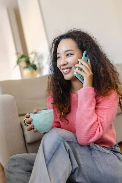 リビングルームでソファに座ってコーヒーを飲みながら 携帯電話を使って話している素敵なアジアの若い女性のイメージ — ストック写真