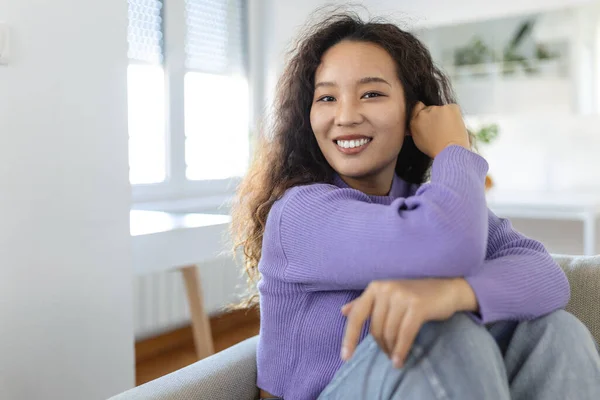 Happy Asian woman sitting on sofa at home and looking at camera. Portrait of comfortable woman in winter clothes relaxing on sofa . Portrait of beautiful woman smiling and relaxing during autumn.