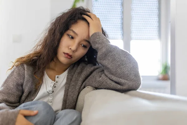 Unhappy Asian woman on sofa crying. Lonely sad woman deep in thoughts sitting daydreaming or waiting for someone in the living room with a serious expression, sitting on couch