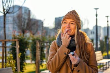 Positive young female in warm coat smiling and looking away while smearing lip balm on lips during stroll on city street