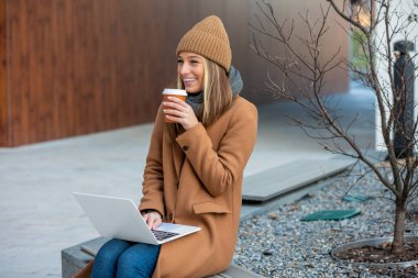 Side view of young woman chatting online on laptop computer, sitting on bench, young female freelancer chatting on social media
