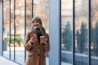 Cheerful young woman wearing coat walking outdoors, holding takeaway coffee cup, using mobile phone