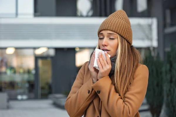 ill beautiful young woman sneezing and blowing nose in napkin. Sick businesswoman in business style sneeze at street. Unhealthy employee. Outdoor. Virus symptoms. Cold disease.
