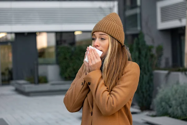 ill beautiful young woman sneezing and blowing nose in napkin. Sick businesswoman in business style sneeze at street. Unhealthy employee. Outdoor. Virus symptoms. Cold disease.