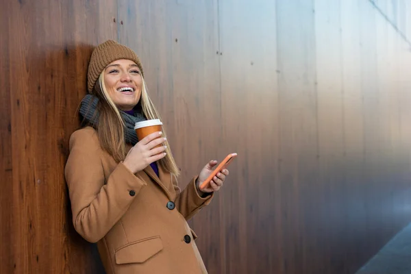 在阳光明媚的城市大街上 身穿外套的快乐的年轻女子用手机 喝着纸杯中的咖啡 — 图库照片