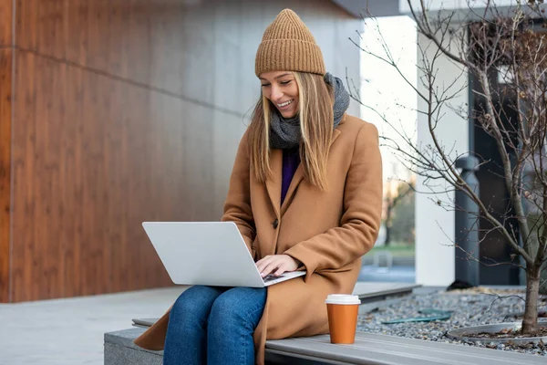 Young woman with wide smile sitting on bench looks at laptop with coffee in her hands on city street. manager in non-office is engaged in additional work at laptop.