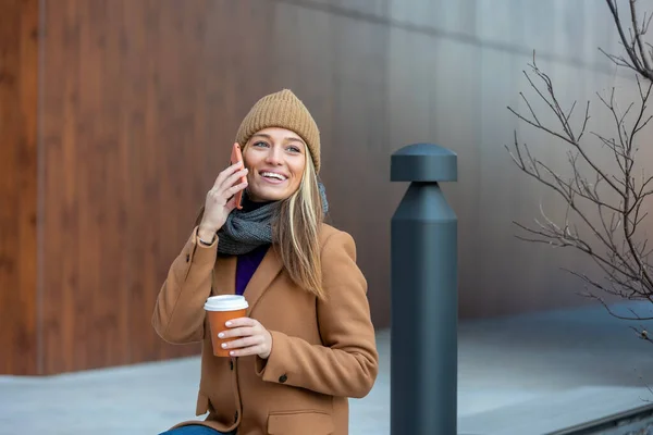 Blonde woman chatting, browsing internet online on smartphone during coffee break while relaxing on park bench. Beautiful cute young woman using mobile phone and holding cup of coffee.