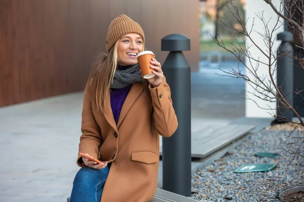 Blonde woman chatting, browsing internet online on smartphone during coffee break while relaxing on park bench. Beautiful cute young woman using mobile phone and holding cup of coffee.