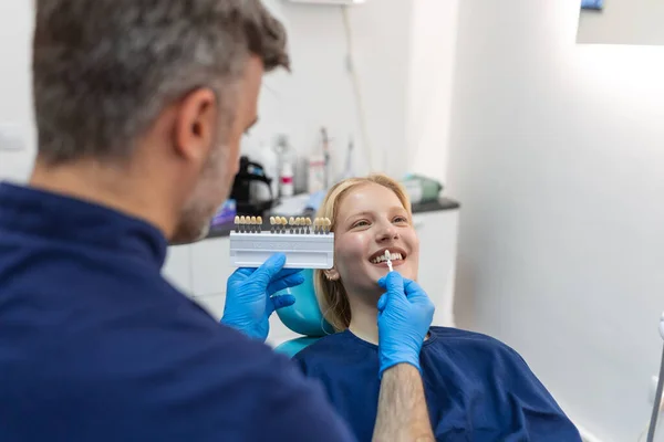 Image of satisfied young woman sitting in dental chair at medical center while professional doctor fixing her teeth, Female dentist choosing filling shade for smiling woman, using tooth scale sample