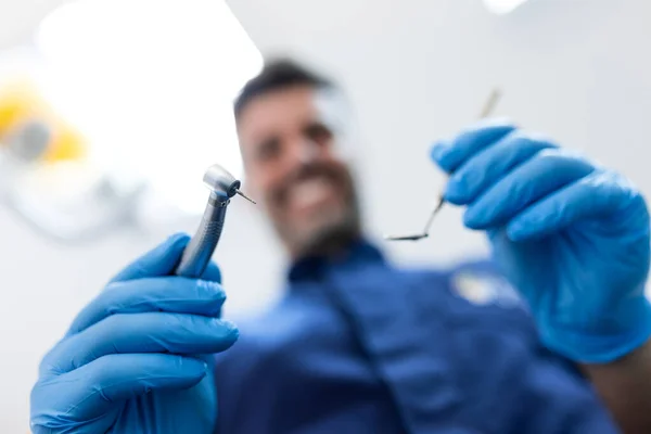 Dental instruments in the hands of a blurred male dentist doing dental examination. Dentist holding angled mirror and dental drill bending over patient