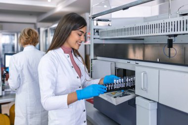 Modern laboratuarda test tüpleri ve analiz makineleriyle analiz yapan genç bir kadın laboratuvar asistanının portresi.