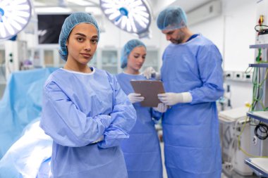 Portrait of female woman nurse surgeon OR staff member dressed in surgical scrubs gown mask and hair net in hospital operating room theater making eye contact smiling pleased happy looking at camera