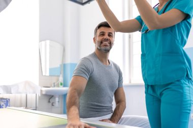 Kadın radyoloji uzmanı hastayı röntgen cihazının yanında el röntgenine hazırlıyor. Radyolog, sedyede yatan hastanın röntgenini çekiyor.