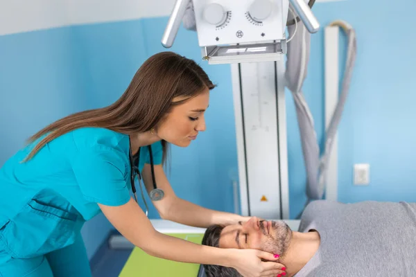 Young confident female doctor radiologist, taking x-ray scan of male patient lying on the machine table. Doctor standing near the patient during head X Ray procedure