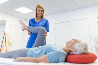 Kadın fizyoterapist, kıdemli yaşlı kadının bacak kaslarını esnetmesine ve egzersiz odasında bacak veya bacak rehabilitasyonuna yardım ediyor. Masaj yatağında yatıyor.