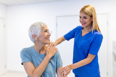 Fizyoterapistle egzersiz yapan mutlu yaşlı kadın. Emekli bir kadın rehabilitasyon sırasında klinikte kişisel antrenörünün yardımıyla kollarını esnetiyor..