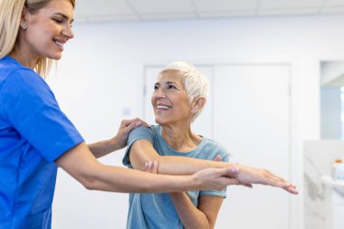 Gün boyunca ofisinde kıdemli bir bayan hastayla çalışan fizyoterapistin fotoğrafı. Fizyoterapist, kas rehabilitasyonu sırasında kadın hastaya yardım ediyor.
