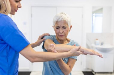Fizyoterapistle egzersiz yapan mutlu yaşlı kadın. Emekli bir kadın rehabilitasyon sırasında klinikte kişisel antrenörünün yardımıyla kollarını esnetiyor..