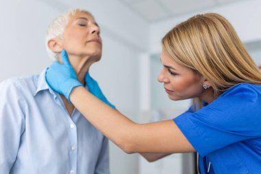 Dost canlısı doktor, eldiven giyip boğaz ve tiroit bezlerini kontrol ediyor, kliniğe gelen yaşlı kadın hastaların boynuna dokunuyor. Tiroid kanseri önleme kavramı